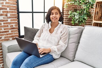 Fototapeta premium Young latin woman using laptop sitting on sofa at home