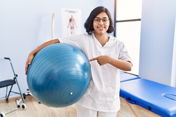 Fototapeta premium Young hispanic physiotherapist woman holding pilates ball smiling happy pointing with hand and finger