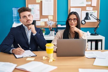 Young hispanic man and woman working at the office serious face thinking about question with hand on chin, thoughtful about confusing idea