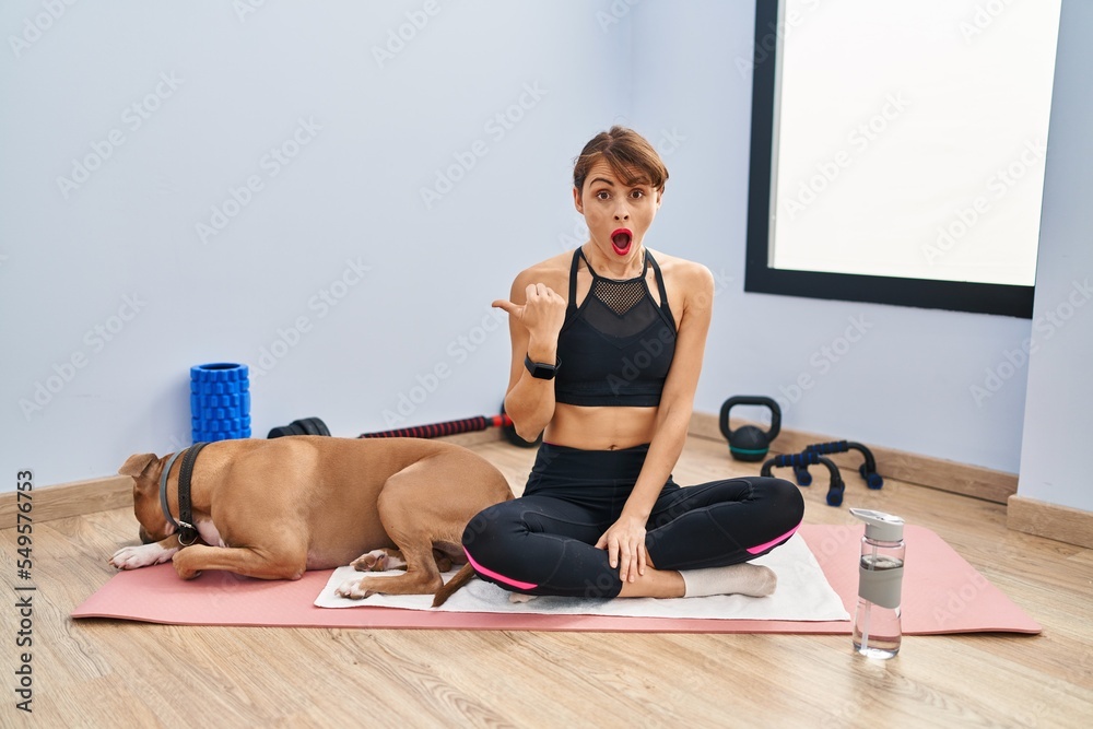 Wall mural Young beautiful woman sitting on yoga mat surprised pointing with hand finger to the side, open mouth amazed expression.