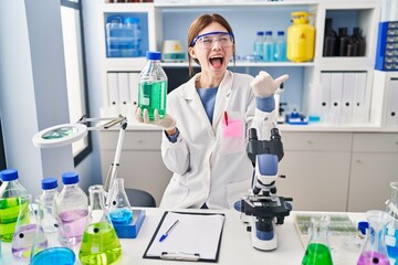 Young brunette woman working at scientist laboratory pointing thumb up to the side smiling happy with open mouth