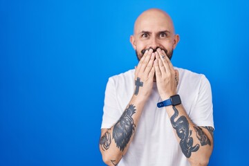 Hispanic man with tattoos standing over blue background laughing and embarrassed giggle covering...