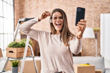 Beautiful woman holding keys of new home showing on video call winking looking at the camera with...