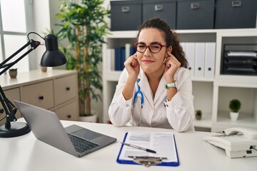 Young hispanic woman wearing doctor uniform and stethoscope covering ears with fingers with annoyed expression for the noise of loud music. deaf concept.