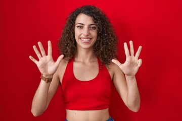 Hispanic woman with curly hair standing over red background showing and pointing up with fingers number nine while smiling confident and happy.