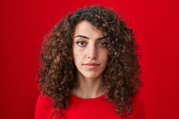 Hispanic woman with curly hair standing over red background relaxed with serious expression on face. simple and natural looking at the camera.