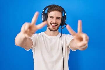 Naklejka premium Hispanic man with beard listening to music wearing headphones smiling with tongue out showing fingers of both hands doing victory sign. number two.