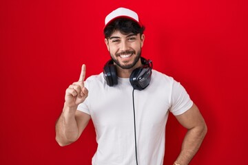 Hispanic man with beard wearing gamer hat and headphones showing and pointing up with finger number one while smiling confident and happy. © Krakenimages.com
