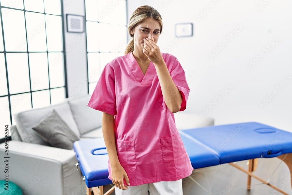 Wall mural young blonde woman working as physiotherapist at home smelling something stinky and disgusting, into