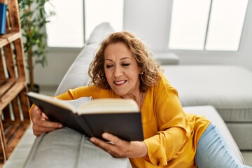 Middle age caucasian woman reading book sitting on the sofa at home.