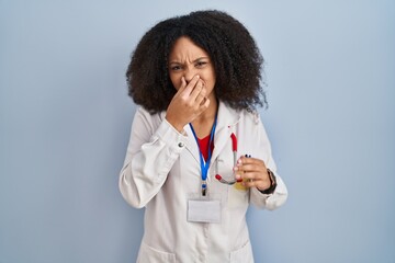 Young african american woman wearing doctor uniform and stethoscope smelling something stinky and disgusting, intolerable smell, holding breath with fingers on nose. bad smell