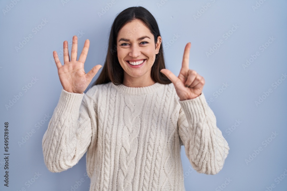 Sticker Young brunette woman standing over blue background showing and pointing up with fingers number seven while smiling confident and happy.