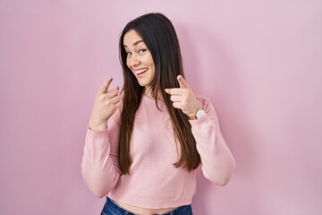 Young brunette woman standing over pink background pointing fingers to camera with happy and funny face. good energy and vibes.
