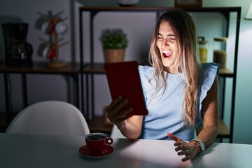 Young hispanic woman using touchpad sitting on the table at night angry and mad screaming frustrated and furious, shouting with anger. rage and aggressive concept.
