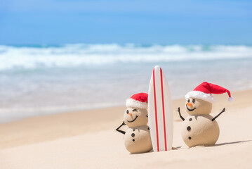 Two Sandy Christmas Snowmen are watching the waves, standing on beautiful beach with a surf board