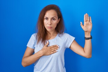 Brunette woman standing over blue background swearing with hand on chest and open palm, making a loyalty promise oath