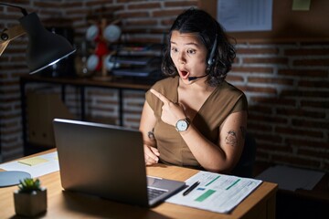 Young hispanic woman working at the office at night surprised pointing with finger to the side, open mouth amazed expression.