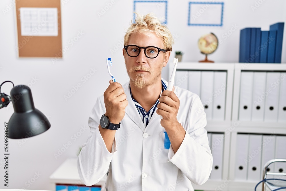 Sticker Young blond man working at dentist clinic holding electric toothbrush and teethbrush skeptic and nervous, frowning upset because of problem. negative person.