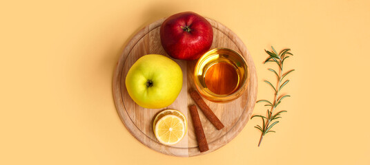 Wooden board with fresh apples, glass of juice, lemon slices and cinnamon sticks on beige background