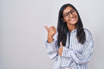 Young hispanic woman wearing glasses pointing to the back behind with hand and thumbs up, smiling confident