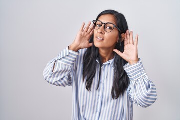 Young hispanic woman wearing glasses trying to hear both hands on ear gesture, curious for gossip. hearing problem, deaf