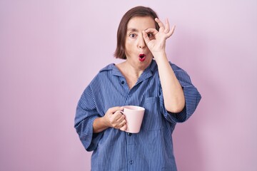 Middle age hispanic woman drinking a cup coffee doing ok gesture shocked with surprised face, eye looking through fingers. unbelieving expression.