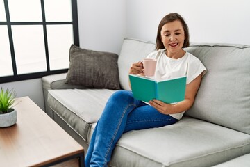 Middle age hispanic woman smiling sitting on the sofa reading a book at home