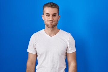 Young caucasian man standing over blue background puffing cheeks with funny face. mouth inflated with air, crazy expression.