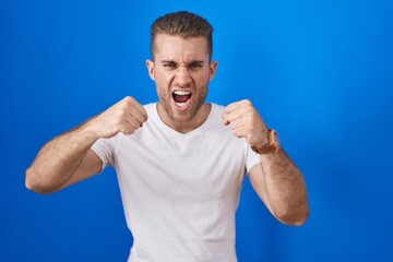 Young caucasian man standing over blue background angry and mad raising fists frustrated and furious while shouting with anger. rage and aggressive concept.