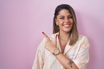 Blonde woman standing over pink background cheerful with a smile of face pointing with hand and finger up to the side with happy and natural expression on face