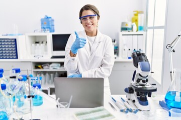 Young hispanic woman wearing scientist uniform working at laboratory smiling happy and positive,...