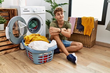 Young caucasian man putting dirty laundry into washing machine smiling looking to the side and staring away thinking.