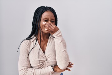 African woman with braids standing over white background smelling something stinky and disgusting, intolerable smell, holding breath with fingers on nose. bad smell - obrazy, fototapety, plakaty