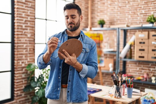 Young hispanic man smiling confident holding paintbrush and palette at art studio