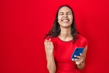 Young brazilian woman using smartphone over red background very happy and excited doing winner...
