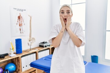 Young caucasian woman working at pain recovery clinic afraid and shocked, surprise and amazed expression with hands on face