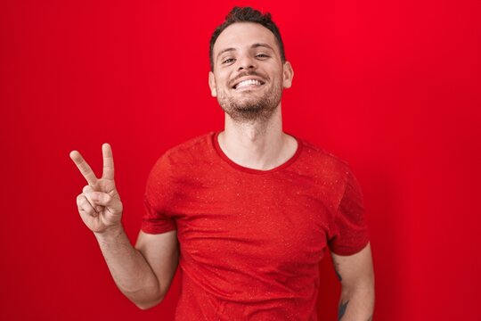 Young hispanic man standing over red background smiling looking to the camera showing fingers doing victory sign. number two.