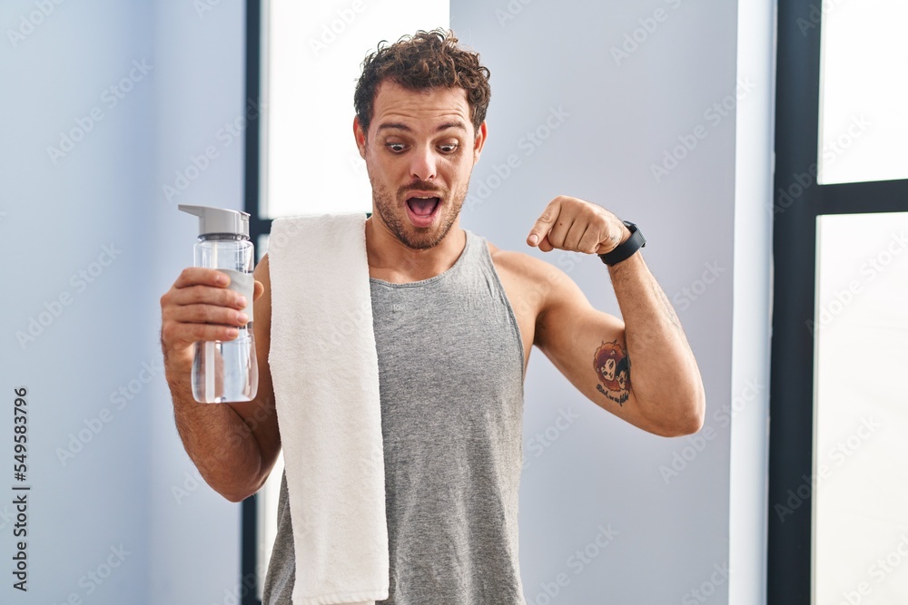 Wall mural Young hispanic man wearing sportswear drinking water pointing down with fingers showing advertisement, surprised face and open mouth