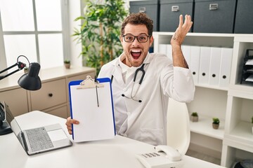Fototapeta premium Young hispanic man wearing doctor stethoscope holding clipboard celebrating victory with happy smile and winner expression with raised hands
