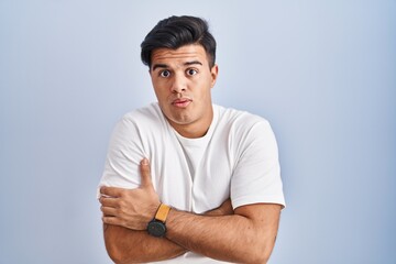 Hispanic man standing over blue background shaking and freezing for winter cold with sad and shock expression on face