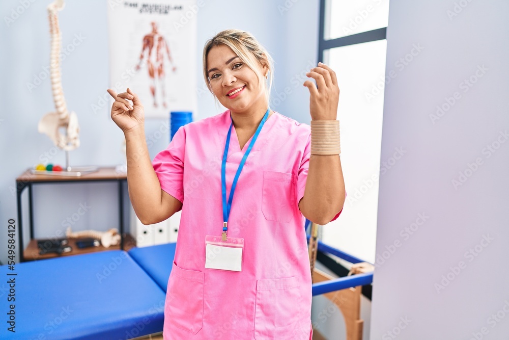 Wall mural young hispanic woman working at pain recovery clinic wearing wristband smiling happy pointing with h