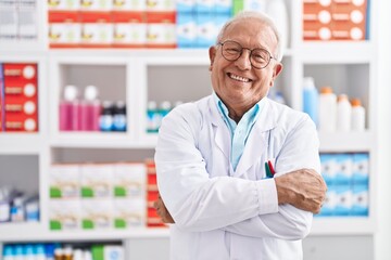 Senior grey-haired man pharmacist smiling confident standing with arms crossed gesture at pharmacy