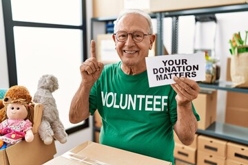 Senior volunteer man holding your donation matters surprised with an idea or question pointing finger with happy face, number one