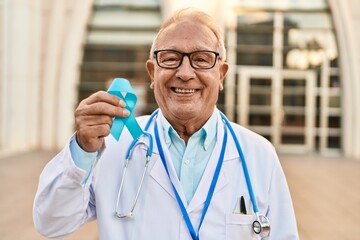 Senior doctor with grey hair holding blue ribbon looking positive and happy standing and smiling...
