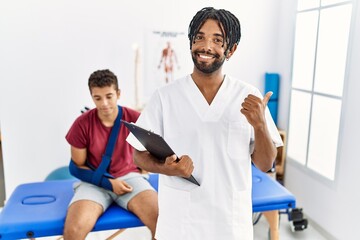 Obraz premium Young hispanic man working at pain recovery clinic with a man with broken arm pointing to the back behind with hand and thumbs up, smiling confident
