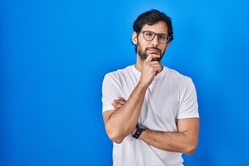 Handsome latin man standing over blue background looking confident at the camera smiling with crossed arms and hand raised on chin. thinking positive.