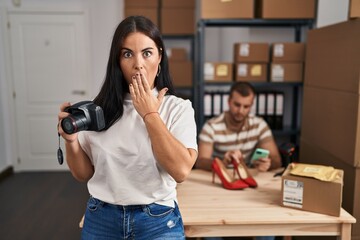 Young hispanic woman holding camera working at small business ecommerce covering mouth with hand,...