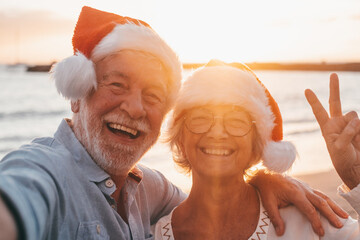 Two old happy seniors wearing christmas hats at the beach taking a selfie of them smiling and...