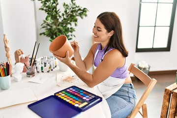 Young beautiful hispanic woman artist smiling confident painting clay pot at art studio