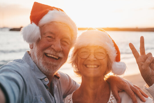 Two old happy seniors wearing christmas hats at the beach taking a selfie of them smiling and having fun with the sunset at the background at evening. Cute couple of old persons looking at the camera